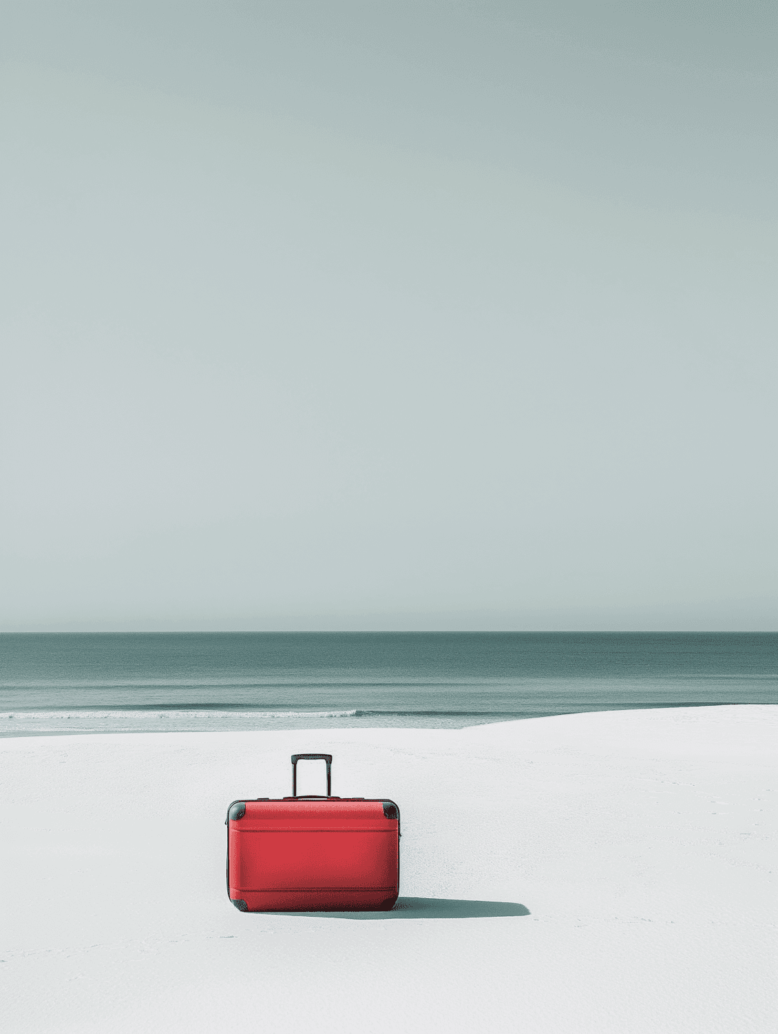 A red suitcase standing on white sand beside a calm sea